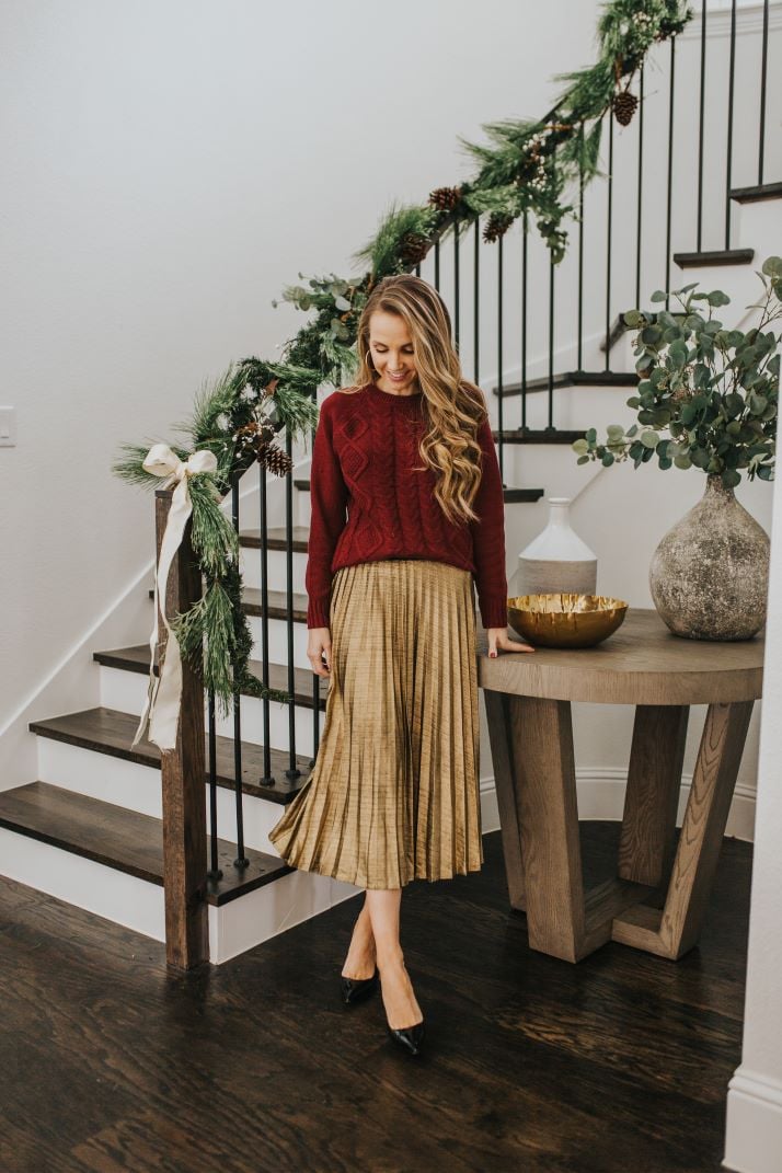 gold pleated skirt and burgundy cable knit sweater in front of curved staircase and round entry table